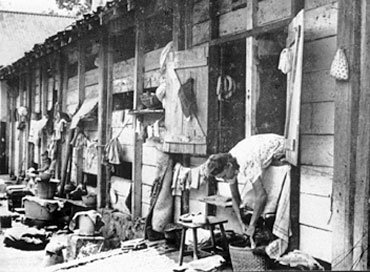 Woman leaning outside a barrack in a row of barracks with laundry and objects in the front.